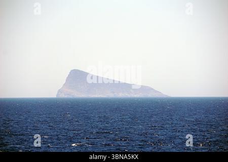 Griechische Inseln zwischen Zypern und Athen. Eine einsame, felsige Insel inmitten eines riesigen, dunkelblauen Ozeans. Die Insel hat eine zerklüftete, felsige Oberfläche und einen kleinen, hellbraunen oder braunen Gipfel. Der Himmel darüber ist hell, blassblau, ohne sichtbare Wolken. Die Perspektive des Bildes ist vom Ozean aus, und blickt auf die Insel. Der Ozean wirkt relativ ruhig mit kleinen Wellen. Das Auge und die Phantasie eines Künstlers Stockfoto