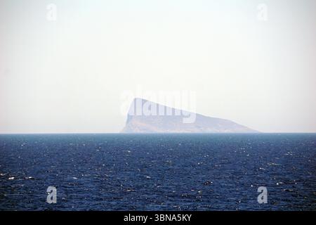 Griechische Inseln zwischen Zypern und Athen. Eine einsame, felsige Insel inmitten eines riesigen, dunkelblauen Ozeans. Die Insel hat eine gedämpfte grau-braune Farbe und eine spitze, leicht geneigte Spitze. Der Himmel darüber ist hell, blassblau und bildet einen Kontrast zu den dunkleren Tönen des Ozeans und der Insel. Die Horizontlinie ist deutlich sichtbar und trennt den Ozean vom Himmel. Das Bild ist aus der Ferne aufgenommen, was ein Gefühl der Isolation und Pracht der Insel vermittelt. Der Ozean wirkt relativ ruhig mit kleinen Wellen. Das Auge und die Phantasie eines Künstlers Stockfoto