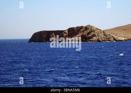 Griechische Inseln zwischen Zypern und Athen. Eine felsige Insel inmitten eines riesigen, tiefblauen Ozeans. Die Insel besteht hauptsächlich aus dunkelbraunen und grauen Felsen, wobei einige hellere Felsen über ihre Oberfläche verstreut sind. Das Meer, das die Insel umgibt, ist relativ ruhig, mit kleinen Wellen, die sanft gegen das felsige Ufer plätschern. Der Himmel darüber ist klar, hellblau. Ein kleines, dunkles Objekt, möglicherweise ein Leuchtturm, ist auf der Spitze der Insel zu sehen. Die Horizontlinie ist in der Entfernung deutlich sichtbar und trennt den Ozean vom Himmel. Das Auge und die Phantasie eines Künstlers Stockfoto