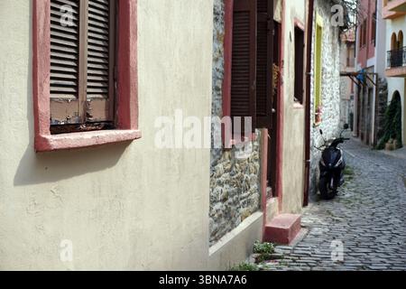 Kavala, Griechenland, Eine enge Kopfsteinpflasterstraße, die von alten Gebäuden mit einer Mischung aus cremefarbenen, beigefarbenen und rosa-roten Wänden flankiert wird. Die Gebäude haben eine Vielzahl von architektonischen Stilen, darunter einige mit Steinmauern und andere mit bemalten oder Stuckwänden. Auf der linken Seite des Bildes ist ein einzelnes Fenster mit einer Holzblende zu sehen, auf der rechten Seite eine geschlossene Holzblende. Ein schwarzer Roller steht auf der rechten Straßenseite. Die kopfsteingepflasterte Straße biegt sich sanft nach rechts und führt zu einem Blick auf ein Gebäude mit einem roten Dach in der Ferne. Das Auge und die Phantasie eines Künstlers Stockfoto