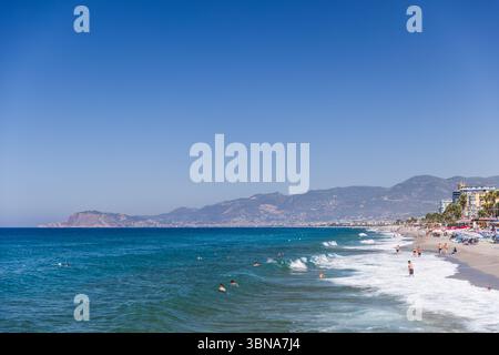 Mahmutlar, Türkei - 13. August 2024: Malerische Küste mit hellblauen Wellen, sonnigem Himmel und Blick auf die Berge. Die Leute schwimmen in Sho Stockfoto