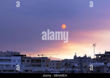 Zypern, Larnaka, das Bild fängt ein Stadtbild bei Sonnenuntergang ein, mit dem die Sonne hinter einer Bergkette in der Ferne untergeht. Der Himmel ist in Orange- und Violetttönen gemalt und schafft eine atemberaubende Kulisse. Das Gebiet ist dicht besiedelt mit Gebäuden, von denen viele Antennen und Satellitenschüsseln auf ihren Dächern haben. Die Gebäude sind überwiegend beige und weiß, einige davon haben einen moderneren architektonischen Stil. Die Stadtlandschaft wird von einem hohen Aussichtspunkt aus betrachtet und bietet einen Panoramablick auf die urbane Landschaft. Die Sonne ist tief am Horizont und strahlt ein warmes Leuchten über die Szene. Ein Baukran Stockfoto