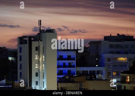 Zypern, Larnaka, das Bild fängt ein Stadtbild in der Abenddämmerung ein, mit einem leuchtenden rosa und orangen Himmel, der die Szene dominiert. Der Himmel ist voller Wolken, die der Atmosphäre Tiefe und Textur verleihen. Das Stadtbild ist eine Mischung aus Gebäuden, mit einem markanten weißen Gebäude auf der linken Seite des Rahmens. Die Gebäude sind mit einer Mischung aus weißem und blauem Licht beleuchtet, was einen Kontrast zu den dunkleren Farben des Himmels schafft. Einige Gebäude verfügen über Balkone und einige Dächer sind mit Antennen und Satellitenschüsseln geschmückt. In der Ferne ist eine Bergkette zu sehen, deren Silhouette sich abhebt Stockfoto