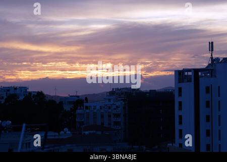 Das Bild fängt eine Skyline bei Sonnenuntergang ein, mit einem leuchtenden Himmel voller Orange-, Rosa- und Lila-Töne. Die Sonne untergeht hinter einer Reihe von Bergen in der Ferne und strahlt ein warmes Licht über die Landschaft. Die Szene ist eine Mischung aus Gebäuden mit einer Vielzahl von architektonischen Stilen und Farben, einschließlich weiß, Grau und Blau. Einige Gebäude haben Antennen und Satellitenschüsseln auf ihren Dächern. Die Skyline wird von einem hohen Aussichtspunkt aus betrachtet und bietet einen Panoramablick auf die Gegend. Das Bild wird aus einer leicht erhöhten Position aufgenommen, sodass Sie einen klaren Blick auf die Skyline haben. Das Auge eines Künstlers und Stockfoto