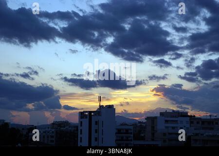 Das Bild fängt ein Stadtbild bei Sonnenuntergang ein, mit einem Himmel voller dunkler, bedrohlicher Wolken. Die Sonne untergeht hinter den Wolken und strahlt ein warmes, oranges und gelbes Leuchten aus. Das Stadtbild bietet eine Mischung aus Gebäuden, darunter ein hohes, weißes Gebäude mit Dachkonstruktion und Antennen, und andere Gebäude mit unterschiedlichen Höhen und Architekturstilen. Die Gebäude zeichnen sich durch eine Silhouette vor dem lebendigen Sonnenuntergangshimmel aus. Die Farben im Bild sind überwiegend Dunkelblau, Schwarz, Orange und Gelb, wobei der Himmel eine Mischung aus dunklen und hellblauen Tönen aufweist. Das Auge und die Phantasie eines Künstlers Stockfoto