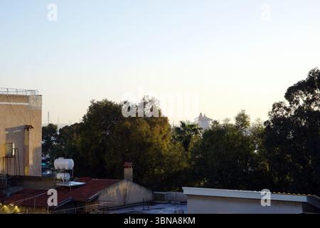 Zypern, Larnaca Salzsee, das Bild erfasst eine Stadtlandschaft von einem Dach aus und bietet einen Panoramablick auf die städtische Landschaft. Der Himmel ist klar, hellblau und die Sonne untergeht und strahlt ein warmes Licht über die Szene. Die Dächer sind mit einer Vielzahl von Gebäuden geschmückt, darunter ein beigefarbenes oder hellbraunes Gebäude mit einem roten Ziegeldach und einem weißen Wassertank an der Seite. Ein weißes Gebäude mit rotem Dach ist ebenfalls in der Mitte des Erdbodens zu sehen. Die Dächer sind mit einer Mischung aus Bäumen und Vegetation bedeckt, einschließlich einer Palme, und ein Teil eines Gebäudes mit einem weißen Dach ist im zu sehen Stockfoto