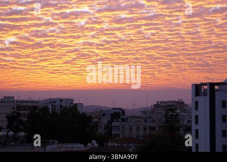 Zypern, Larnaka, Eine Stadtlandschaft bei Sonnenuntergang, mit einem leuchtenden orange-rosa Himmel, der den oberen Teil dominiert. Der Himmel ist mit einer Vielzahl von Wolken gefüllt, die ein strukturiertes und mehrschichtiges Erscheinungsbild schaffen. In der Ferne stehen mehrere Windräder hoch vor dem Hintergrund der Gebäude. Die Szene selbst ist eine Mischung aus Strukturen mit einer Vielzahl von architektonischen Stilen und Farben, einschließlich weiß, Beige und Grau. Einige Gebäude verfügen über Dachkonstruktionen oder Antennen. Die Stadtlandschaft ist von Bäumen und Vegetation umgeben und verleiht der Stadtlandschaft einen Hauch von Natur. Das Bild wurde aus einem hohen Blickwinkel aufgenommen Stockfoto