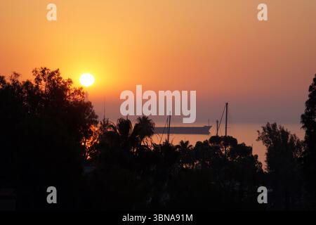 Ein atemberaubender Sonnenuntergang über einem Gewässer, mit Sonnenuntergang hinter einer Reihe von Bäumen auf der linken Seite. Der Himmel ist in Orange- und Rosa-Tönen gemalt und bildet eine atemberaubende Kulisse für die untergehende Sonne. Die Sonne befindet sich auf der linken Seite des Bildes und strahlt ein warmes Leuchten über die Szene aus. In der Ferne ist ein großes Schiff auf dem Wasser zu sehen, das vor dem pulsierenden Himmel eine Silhouette bildet. Das Wasser wirkt ruhig, und die Bäume im Vordergrund sind dunkle Silhouetten vor dem hellen Himmel. Ein Künstlerauge und fantasievolle Bildunterschrift. Zypern, Larnaka Stockfoto