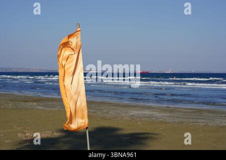 Zypern, Larnaka, Eine ruhige Strandszene. Im Vordergrund steht eine einsame orange Flagge auf einem Metallmast, deren leuchtende Farbe sich mit dem hellbraunen Sand abhebt. Die Flagge weht leicht im Wind und verleiht der ansonsten ruhigen Umgebung ein Gefühl von Bewegung. Jenseits der Flagge erstreckt sich die Weite des Ozeans, dessen Oberfläche von kleinen Wellen gespickt ist. Das Wasser erscheint dunkelblau-grün. In der Entfernung ist ein rotes Boot am Horizont zu sehen, und eine kleine, undeutliche Struktur ist auch am Horizont zu sehen. Der Himmel ist klar, hellblau, und der Sand scheint ein Licht zu sein Stockfoto