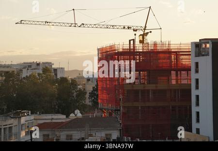 Zypern, Larnaka, Eine geschäftige Baustelle bei Sonnenuntergang. Ein hoch aufragender Kran mit einem langen Arm und einem gelben Eimer am Ende steht vor dem Hintergrund einer Skyline der Stadt. Der Arm des Krans wird ausgefahren und reicht in Richtung Himmel, und die Schaufel befindet sich in der Nähe der Oberseite des Krans. Die Baustelle ist teilweise durch ein rotes Netz oder Gerüst umschlossen, und ein im Bau befindliches Gebäude ist im Vordergrund sichtbar. Der Himmel ist in Orange- und Rosa-Tönen gemalt und spiegelt den warmen Glanz der untergehenden Sonne wider. Das Stadtbild im Hintergrund zeigt eine Mischung von Gebäuden, darunter ein weißes Gebäude Stockfoto