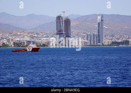 Limassol, Zypern, Eine pulsierende Landschaft vor dem Hintergrund majestätischer Berge. Die Szene ist eine Mischung aus modernen Hochhäusern und älteren Bauwerken, wobei auf einem der Gebäude ein prominenter Baukran steht. Das Gebiet verfügt über tiefblaues Wasser, das sich bis zum Horizont erstreckt. Ein rot-weißes Schlepper ist im Wasser zu sehen, und einige kleine Boote können in der Entfernung gesehen werden. Die Berge im Hintergrund sind eine Mischung aus braunen und braunen Farben, wobei einige Bereiche trocken oder karg wirken. Der Himmel ist klar, hellblau. Das Auge und die Phantasie eines Künstlers Stockfoto