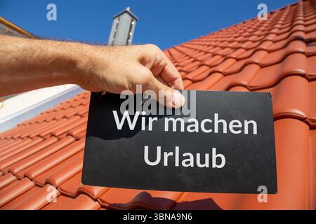 Dachdecker Firmenfeiertage: Mann mit Schild (mit hinzugefügtem Text) wir MACHEN URLAUB vor einem neuen Ziegeldach Stockfoto