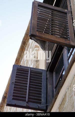 Nikosia (Lefkosia), Zypern, die letzte geteilte Hauptstadt der Welt, Ein Teil eines Gebäudes mit einer Steinfassade und einem hölzernen Fensterrahmen. Das Fenster verfügt über zwei dunkelbraune Holzläden, von denen einer teilweise offen ist und einen Blick auf das Innere freigibt. Die Rollläden sind mit Metallhalterungen am Fensterrahmen befestigt. Das Fenster ist in eine Steinmauer eingefasst, die eine hellbeige oder cremefarbene Farbe hat. Der Himmel ist klar, hellblau. Das Auge und die Phantasie eines Künstlers Stockfoto