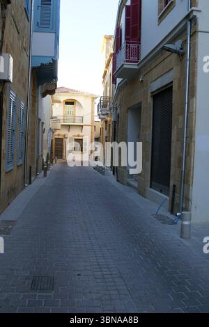 Nikosia (Lefkosia), Zypern, die letzte geteilte Hauptstadt der Welt, Eine enge Kopfsteinpflasterstraße in mediterranem Stil. Die Straße wird von Gebäuden aus hellem Stein flankiert, einige mit roten Fensterläden. Die Gebäude sind überwiegend beige oder cremefarbene Gebäude, einige haben Balkone. Auf der linken Straßenseite ist ein blau gestrichenes Gebäude zu sehen. Die Straße ist frei von Menschen und Fahrzeugen, was ein Gefühl der Ruhe schafft. Der Himmel ist klar, hellblau und die Straße scheint relativ leer zu sein, ohne sichtbare Anzeichen von Aktivität. Ein Stockfoto