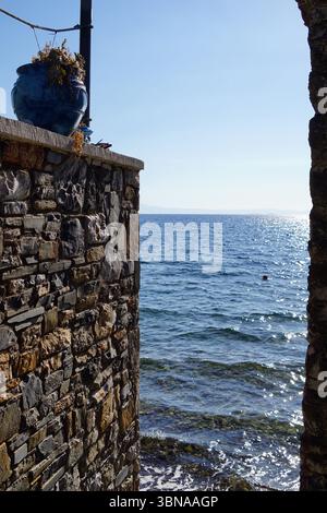NIEs, Griechenland, Eine ruhige Küstenszene, von einer Steinmauer aus gesehen, die den Blick umrahmt. Die Wand, die aus unregelmäßig geformten Steinen in Braun-, Grau- und Beigetönen besteht, ist mit einem blauen Topf mit einer Pflanze geschmückt, was der Umgebung einen Hauch von Natur verleiht. Der Topf befindet sich auf einem kleinen Vorsprung oder einer Plattform an der Wand. Jenseits der Mauer erstreckt sich die Weite des tiefblauen Ozeans, mit kleinen Wellen, die sanft gegen das Ufer plätschern. Der Himmel darüber ist klar, hellblau, und eine kleine rote Boje ist im Wasser zu sehen. Die Perspektive des Bildes ist aus einem niedrigen Blickwinkel, der nach oben blickt Stockfoto