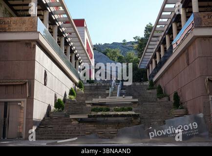 Eine große Treppe mit einem Metallgeländer, die zu einem Hügel hinaufsteigt. Das Treppenhaus wird von zwei Gebäuden flankiert, eines mit einer roten und das andere mit einer braunen Fassade. Die Gebäude verfügen über Balkone mit weißen Geländern und Markisen. Die Treppe ist mit kleinen Pflanzen und Blumen geschmückt, die der städtischen Umgebung einen Hauch von Grün verleihen. Der Hügel im Hintergrund ist mit Bäumen und Sträuchern bedeckt, was einen natürlichen Kontrast zu den künstlichen Strukturen bildet. Der Himmel ist klar blau, und die gesamte Szene ist in natürliches Licht getaucht. Im bo ist ein Schild mit der Aufschrift "KULTURHAUPTSTADT EUROPAS" zu sehen Stockfoto
