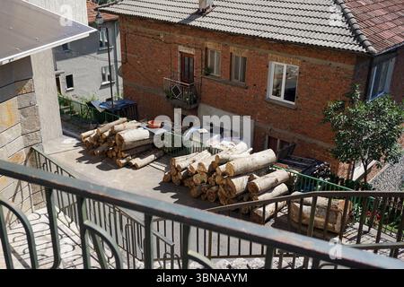 Smolyan, Bulgarien, Ein Backsteingebäude mit grauem Ziegeldach. Vor dem Gebäude befindet sich ein großer Stapel ordentlich gestapelter Baumstämme mit einem kleinen Tisch und einem roten Eimer in der Nähe. Ein grünes Geländer verläuft am Rand des gepflasterten Bereichs vor dem Gebäude entlang. Ein kleiner Balkon mit Geländer ist im zweiten Stock des Gebäudes zu sehen. Rechts vom Gebäude ist ein kleiner Baum zu sehen. Ein Teil einer Stein- oder Betonwand ist auf der linken Seite des Bildes sichtbar. Die Szene wird von einem hohen Aussichtspunkt aus betrachtet und bietet einen umfassenden Blick auf das Gebäude und seine Umgebung. Das Auge eines Künstlers und Stockfoto