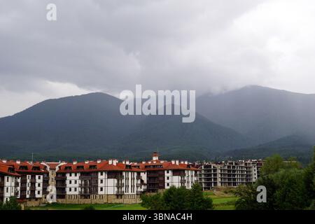 Eine ruhige Landschaft mit einer Ansammlung von weißen und roten Gebäuden in einem Tal, umgeben von üppigen grünen Bäumen. Die Gebäude, die wie Wohn- oder Ferienobjekte wirken, liegen leicht abschüssig und sind teilweise von der umgebenden Vegetation verdeckt. Der Himmel ist bedeckt mit grauen Wolken, die ein weiches Licht über die Szene werfen. Im Hintergrund erstreckt sich eine majestätische Bergkette über den Horizont, deren Gipfel in Nebel oder Nebel gehüllt sind. Die Berge sind von dichtem Wald bedeckt, und die Farbpalette des Bildes wird von Grün-, Grau- und Weißtönen dominiert. A Stockfoto