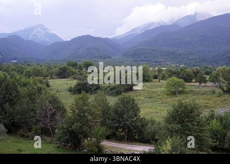 Eine ruhige Landschaft mit einem üppig grünen Feld mit Bäumen und Büschen. Im Vordergrund ist eine unbefestigte Straße oder ein Pfad zu sehen, die in Richtung des Feldes führt. In der Ferne erheben sich majestätische Berge, deren Gipfel von einer Mischung aus grüner und brauner Vegetation bedeckt sind, mit einigen schneebedeckten Gebieten. Der Himmel ist bedeckt und hat eine hellgraue oder weiße Farbe. Das Bild wird aus einem hohen Blickwinkel aufgenommen und bietet einen Panoramablick auf die Landschaft. Bansko ist eine malerische Stadt im Südwesten Bulgariens, eingebettet am Fuße des Pirin-Gebirges. Stockfoto
