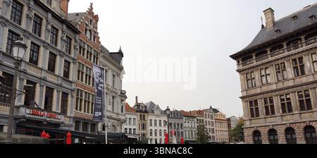 Antwerpen, zweitgrößte belgische Stadt an der Schelde in Belgien, Ein kopfsteingepflasterter Platz, wahrscheinlich ein historisches Zentrum. Der Platz ist von einer Vielzahl von Gebäuden umgeben, einschließlich einer Mischung aus architektonischen Stilen, von denen einige mit kunstvollen Details und andere mit einfacheren Fassaden ausgestattet sind. Die Gebäude sind hauptsächlich in Beige-, Grau- und Weißtönen gehalten, mit einigen roten Akzenten. Der Platz ist voller Menschen, die in Cafés im Freien laufen und sitzen, die mit Tischen und Stühlen eingerichtet sind. Einige rote Regenschirme sind ebenfalls zu sehen, was der Szene einen Farbakzent verleiht. Der Himmel ist bewölkt und wirft einen Stockfoto