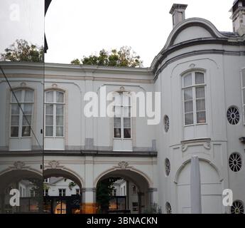 Ein Innenhof mit Kopfsteinpflasteroberfläche, umgeben von einem weißen Gebäude mit Bogenfenstern und einer dekorativen Fassade. Das Gebäude verfügt über einen großen, bogenförmigen Eingang und eine weiße Tür. Im Innenhof sind mehrere Tische und Stühle angeordnet, einige mit weißen Tischdecken und andere mit dunklen Tischdecken. Ein paar Topfpflanzen sind um den Innenhof herum platziert, was einen Hauch von Grün verleiht. Ein paar Leute gehen durch den Innenhof und ein weißer Schirm ist in der Nähe des Gebäudes zu sehen. Der Innenhof ist von Bäumen eingerahmt und der Himmel ist bedeckt. Antwerpen, zweitgrößte belgische Stadt, liegt an der S Stockfoto