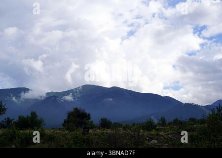 Eine majestätische Bergkette mit einer Mischung aus grüner und brauner Vegetation, einschließlich Bäumen und Sträuchern. Die Berge sind von einer Wolkenschicht bedeckt, wobei einige Bereiche dunkler und schattiger erscheinen. Der Himmel ist hellblau mit verstreuten weißen Wolken. Der Vordergrund zeigt eine Mischung aus grüner und brauner Vegetation, wobei einige Felsen und kleine Sträucher sichtbar sind. Die Berge im Hintergrund sind dunkler grün, die Gipfel sind teilweise von Wolken verdeckt. Bansko ist eine malerische Stadt im Südwesten Bulgariens, eingebettet am Fuße des Pirin Moun Stockfoto