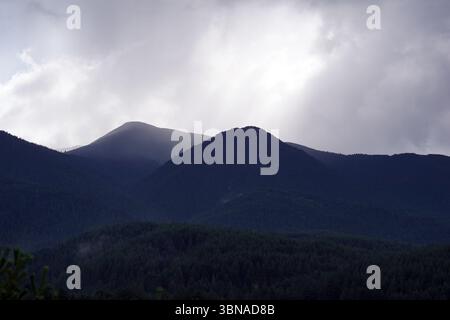 Eine majestätische Bergkette, umgeben von einer dicken Wolkenschicht. Die Berge, in einem dunkelgrünen Farbton, erheben sich majestätisch gegen den Himmel. Die Gipfel werden teilweise von den Wolken verdeckt, was ein Gefühl von Tiefe und Geheimnis schafft. Die Berge sind von einem dichten Wald aus dunkelgrünen Bäumen bedeckt, die wie Nadelbäume aussehen. Der Himmel ist hellgrau oder weiß, mit Sonnenlichtern, die durch die Wolken brechen und ein sanftes Leuchten auf die Berge werfen. Bansko ist eine malerische Stadt im Südwesten Bulgariens, eingebettet am Fuße des Pirin-Gebirges. Stockfoto