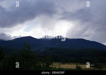 Eine majestätische Bergkette, umgeben von einer dicken Wolkenschicht. Die Berge, bedeckt mit einer Mischung aus dunkelgrüner und brauner Vegetation, dominieren die Szene. Der Himmel ist eine Leinwand aus grauen und weißen Wolken, durch die blaue Flecken hindurchblicken. Die Berge scheinen sich in einem Tal oder tief gelegenen Gebiet zu befinden, mit einem Feld aus trockenem, goldbraunem Gras im Vordergrund. Das Bild ist aus einem niedrigen Winkel aufgenommen und unterstreicht die Pracht der Berge und die Weite des Himmels. Die Berge sind nicht gerade, manche zeigen eine leichte Wellenbewegung. Ein Künstlerauge und fantasievolle Bildunterschrift. Bansko ist A Stockfoto