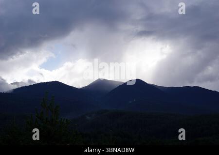 Eine majestätische Bergkette, umgeben von einer dicken Wolkenschicht. Die Berge erheben sich majestätisch gegen den Himmel, deren Gipfel teilweise von den Wolken verdeckt sind. Die Berge sind hauptsächlich dunkelgrün, einige Bereiche sind fast schwarz. Der Himmel ist eine Mischung aus dunkelgrauen und weißen Wolken, mit sichtbaren blauen Flecken. Die Berge sind von einem bewaldeten Gebiet mit einer Mischung aus dunkelgrünen und braunen Bäumen umgeben. Das Bild wird von einem hohen Aussichtspunkt aufgenommen und bietet einen Panoramablick auf die Berge und den umliegenden Wald. Ein Auge und eine fantasievolle Bildunterschrift., B Stockfoto
