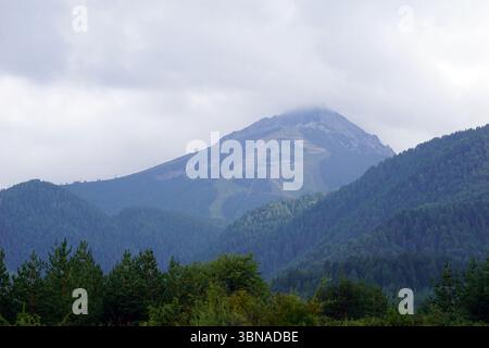 Eine majestätische Bergkette mit einem markanten Gipfel, der von einer Wolkenschicht umhüllt ist. Die Hänge des Berges sind von einem dichten Wald aus dunkelgrünen Bäumen bedeckt, mit Anklängen von hellgrünem und braunem Laub. An den Hängen des Berges ist ein Skigebiet oder eine ähnliche Entwicklung zu sehen. Der Himmel ist bedeckt mit einer Mischung aus grauen und weißen Wolken. Die Perspektive des Bildes ist von einem niedrigeren Aussichtspunkt aus, der auf den Berg blickt und dessen Pracht und Tiefe der bewaldeten Hänge betont. Bansko ist eine malerische Stadt im Südwesten Bulgariens, eingebettet in Th Stockfoto