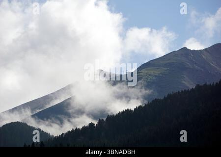 Eine majestätische Bergkette mit einer Mischung aus Grün- und Brauntönen. Die Berge sind teilweise von einer dicken Schicht weißer Wolken verdeckt, was eine dramatische und stimmungsvolle Atmosphäre schafft. Die Berge sind von einer Mischung aus grüner und brauner Vegetation bedeckt, wobei einige Gebiete karger erscheinen als andere. Im Vordergrund erstreckt sich ein dichter Wald aus dunkelgrünen Bäumen über den unteren Teil des Bildes, der einen starken Kontrast zu den helleren Tönen der Berge bildet. Der Himmel darüber ist ein klares Blau mit flauschigen weißen Wolken. Bansko ist eine malerische Stadt, die das Auge und die Fantasie eines Künstlers darstellt Stockfoto