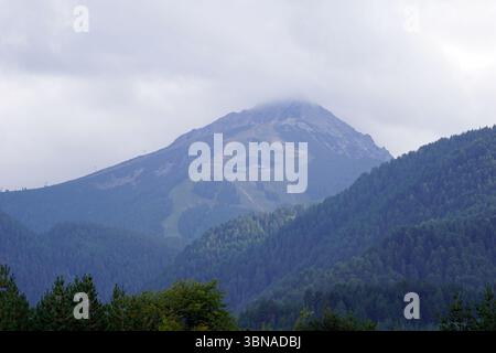 Eine majestätische Bergkette, umgeben von einer dicken Nebelschicht. Der Berg mit seinem zerklüfteten, felsigen Gipfel dominiert die Szene. Die Hänge sind von einer Mischung aus grüner und brauner Vegetation bedeckt, wobei einige Bereiche scheinbar von Bäumen befreit sind. Der Gipfel des Berges ist teilweise durch den Nebel verdeckt, was ihm ein trübes Aussehen verleiht. Der Himmel ist bedeckt und hat eine hellgraue oder weiße Farbe. Im Vordergrund bildet ein Wald aus dunkelgrünen Bäumen einen Kontrast zur zerklüfteten Landschaft des Berges. Bansko ist eine malerische Stadt im Südwesten Bulgariens, eingebettet am Fuße des Künstlers Stockfoto