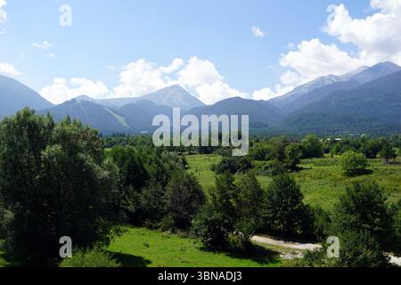 Eine ruhige Landschaft mit einem üppigen grünen Feld, das sich bis zu einer Reihe majestätischer Berge erstreckt. Die Berge, deren Gipfel sich zum klaren blauen Himmel hin erstrecken, sind von einer Mischung aus grüner und brauner Vegetation bedeckt. Das Feld ist mit Bäumen und Büschen gespickt, was der Szene einen Hauch von Leben verleiht. Im Vordergrund ist eine Schotterstraße zu sehen, die den Blick auf die Berge führt. Der Himmel ist ein klares Blau mit einigen verstreuten weißen Wolken. Bansko ist eine malerische Stadt im Südwesten Bulgariens, eingebettet am Fuße des Pirin-Berges Stockfoto