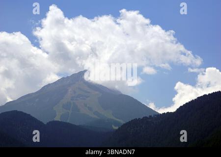 Eine majestätische Bergkette unter einem klaren blauen Himmel. Der Berg, dessen Gipfel in Richtung Himmel reicht, ist von üppiger grüner Vegetation bedeckt. An den Hängen des Berges ist ein Pfad oder Pfad zu sehen. Der Berg ist von einer Mischung aus dunkelgrünen und braun/schwarzen Bäumen umgeben, die einen Kontrast zum blauen Himmel und dem Gipfel des Berges bilden. Der Gipfel des Berges ist teilweise von einer großen, flauschigen weißen Wolke verdeckt. Bansko ist eine malerische Stadt im Südwesten Bulgariens, eingebettet am Fuße des Pirin-Gebirges. Stockfoto