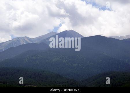 Eine majestätische Bergkette mit einer Mischung aus grünen und braunen/grauen Bergen. Die Berge sind von einem dichten Wald aus dunkelgrünen Bäumen bedeckt, wobei einige Bereiche hellgrün wirken. Der Himmel ist hellblau mit flauschigen weißen Wolken. Die Perspektive des Bildes ist aus einem niedrigen Winkel, der auf die majestätischen Berge blickt und ein Gefühl von Größe und Größe schafft. Das Bild ist frei von Text oder von Menschen geschaffenen Objekten und konzentriert sich ausschließlich auf die natürliche Landschaft. Bansko ist eine malerische Stadt im Südwesten Bulgariens, eingebettet am Fuße des Pirinbergs Stockfoto