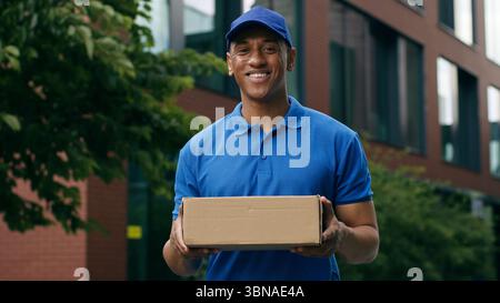 Lächelndes afroamerikanisches ethnisches Kurierporträt, das in der Stadt steht, Lächeln, Kamerazustellmann, männlicher Postarbeiter, der Paketpost hält Stockfoto