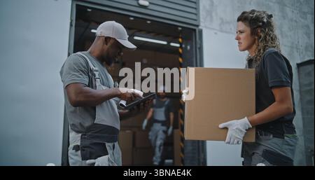 Verschiedene Mitarbeiter, die Kartons zum Logistiklager transportieren. Afroamerikaner-Koordinator scannt Pakete mit Barcode-Scanner mit digitalem Tablet. Online-Bestellungen, Lieferung, E-Commerce-Waren. Stockfoto