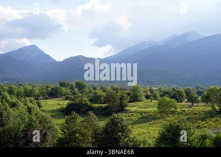 Bansko ist eine malerische Stadt im Südwesten Bulgariens, eingebettet am Fuße des Piringebirges. Eine ruhige Landschaft mit einem üppigen grünen Feld im Vordergrund, gespickt mit Bäumen und Büschen. Jenseits des Feldes erstreckt sich eine Reihe majestätischer Berge am Horizont, deren Gipfel von einem hellblauen Himmel mit verstreuten weißen Wolken umhüllt sind. Die Berge sind hauptsächlich von dunkelgrüner Vegetation bedeckt, wobei einige Bereiche Braun oder Grau aufweisen. Die Berge scheinen eine Mischung aus verschiedenen Arten von Vegetation zu sein, einschließlich Bäumen und Sträuchern. In der Mitte befindet sich ein kleines Gebäude oder Gebäude Stockfoto