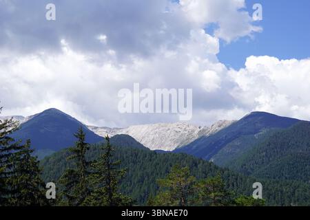 Eine majestätische Bergkette unter einem klaren blauen Himmel mit flauschigen weißen Wolken. Die Berge mit ihren unterschiedlichen Grün- und Brauntönen sind von einer Mischung aus Bäumen und Sträuchern bedeckt. Die Gipfel der Berge sind sichtbar, einige davon sind bläulich-grau gefärbt. Die Berge sind hauptsächlich mit Bäumen bedeckt, wobei einige Bereiche eine Mischung aus Grün und Braun aufweisen. Das Bild wird von einem hohen Aussichtspunkt aufgenommen und bietet einen Panoramablick auf die Berge und die umliegende Landschaft. Bansko ist eine malerische Stadt im Südwesten Bulgariens, am Fuße von t Stockfoto