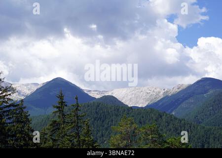 Eine majestätische Bergkette unter bewölktem Himmel. Die Berge mit ihren Gipfeln, die sich in Richtung Himmel erstrecken, sind von einer Mischung aus grüner und brauner Vegetation bedeckt, darunter immergrüne Bäume und einige Bereiche mit Schneeflächen. Die Berge sind hauptsächlich von grüner Vegetation bedeckt, wobei einige Bereiche braune oder graue Flecken aufweisen. Der Himmel ist eine Mischung aus Blau und weiß, mit flauschigen weißen Wolken, die darüber verstreut sind. Das Bild wurde von einem hohen Aussichtspunkt aufgenommen und bietet einen Panoramablick auf die Berge und die umliegende Landschaft. Bansko ist eine malerische Bildunterschrift mit Künstleraugen und Fantasie Stockfoto