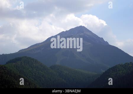 Eine majestätische Bergkette mit einem markanten Gipfel im Zentrum. Der Berg ist von einer Mischung aus grüner und brauner Vegetation bedeckt, wobei einige Bereiche dunkler erscheinen, möglicherweise aufgrund von Schatten oder der Art der Vegetation. Der Gipfel hat eine robuste, felsige Oberfläche. Der Himmel darüber ist hellblau, mit flauschigen weißen Wolken gesäumt. Die Perspektive des Bildes ist von einem unteren Blickpunkt aus, der auf den Berg blickt und ein Gefühl von Größe und Größe schafft. Die Bergkette liegt eingebettet in ein bewaldetes Gebiet, mit Bäumen, die dunkelgrün und üppig erscheinen. Bansko ist eine malerische Stadt im Südwesten Bulgariens Stockfoto