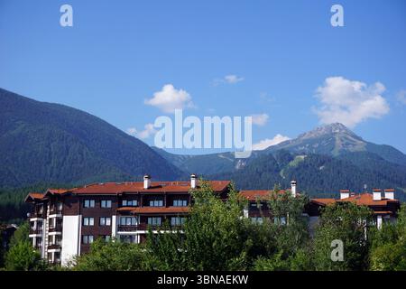 Ein großes, mehrstöckiges Gebäude mit einem roten Dach und weißen Wänden. Das Gebäude hat mehrere Kamine und ist von üppigem Grün umgeben, darunter Bäume und Büsche. Im Hintergrund erstreckt sich eine majestätische Bergkette über den Horizont, deren Gipfel bis zum klaren blauen Himmel reichen. Die Berge sind von einer dichten Schicht von Bäumen und Vegetation bedeckt. Der Himmel ist hellblau mit ein paar verstreuten weißen Wolken. Bildunterschrift in Form eines Künstlerauges und einer phantasievollen Bildunterschrift. Bulgarien, Bansko, Pirinberg Stockfoto
