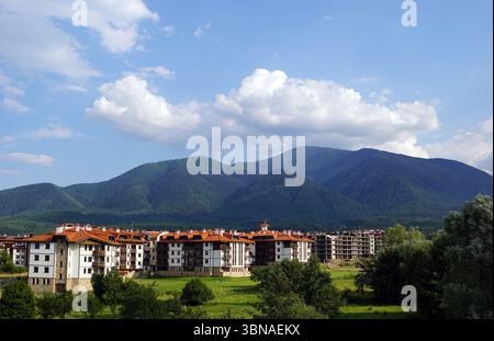 Eine ruhige Landschaft mit einer Ansammlung von weißen und braunen Gebäuden in einem grünen Tal. Die Gebäude, die wie Wohn- oder Ferienhäuser aussehen, sind etwas linear angeordnet, wobei einige Gebäude näher im Vordergrund und andere weiter hinten liegen. Die Gebäude haben rote Ziegeldächer, die der Szene einen Farbakzent verleihen. Das Tal ist von majestätischen, dunkelgrünen Bergen umgeben, deren Gipfel bis zum klaren blauen Himmel reichen. Die Berge sind mit einer dichten Baumschicht bedeckt, die eine üppige grüne Kulisse schafft. Der Himmel ist hellblau, mit flauschigen weißen Wolken gesäumt. Die Stockfoto