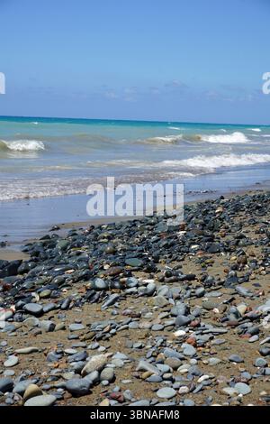 Cyprus Beach, Ein felsiger Strand mit einer Mischung aus dunklen und hellgrauen/blauen Steinen, verstreut am Sandstrand. Der Strand erstreckt sich zum Wasser hin, wo das Wasser eine hellblau-grüne Farbe hat und die Wellen auf das Ufer stürzen. Der Himmel ist klar, hellblau, und der Horizont ist in der Ferne sichtbar. Die Perspektive des Bildes ist vom Strand aus, blickt auf das Wasser und erzeugt ein Gefühl von Tiefe und Stockfoto