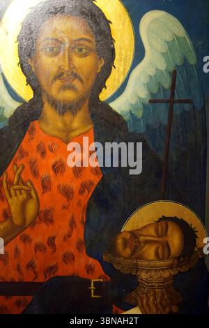 Eine religiöse Ikone, möglicherweise ein Gemälde oder eine Holzschnitzerei, mit einer zentralen Figur mit dunklen Haaren und einem Bart, die ein leuchtendes rot-schwarz gemustertes Kleidungsstück trägt. Das Gesicht der Figur ist teilweise von einem goldenen Halo verdeckt, und ihre Hände werden in einer Geste erhoben, die als Segen oder Friedensgeste interpretiert werden könnte. Die Figur ist von einem blauen Hintergrund mit einem goldenen Halo umgeben, und hinter der zentralen Figur befindet sich ein weißer Engel mit ausgestreckten Flügeln. Ein Kreuz ist auf einem Ständer hinter dem Engel sichtbar. In der unteren rechten Ecke ein Teil eines Gesichts, möglicherweise einer bedeutenden Religion Stockfoto