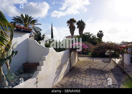 Eine ruhige Gartenszene mit einer weißen Stuckwand und einem Terrakotta-Dach auf der linken Seite des Rahmens. Die Wand ist mit einer Vielzahl von Pflanzen geschmückt, darunter ein Kakteen, ein kleiner Baum und ein blühender Busch mit rosa Blüten. Ein Kiesweg führt zu einem kleinen Gebäude im Hintergrund, das wie ein Haus aussieht. Der Garten ist von üppigem Grün umgeben, einschließlich Palmen und anderen tropischen Pflanzen, die eine ruhige Atmosphäre schaffen. Der Himmel ist klar blau mit flauschigen weißen Wolken, und die Sonne scheint hell und wirft Schatten auf den Boden. Eine kleine, dunkle Struktur, Stockfoto