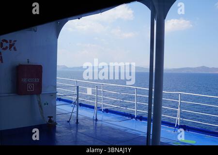 Griechische Inseln zwischen Zypern und Athen., Ein Blick vom Deck eines Schiffes, Blick auf die weite Weite des Ozeans. Die Terrasse ist in einem auffälligen Blauton gestrichen und verfügt über ein weißes Geländer, das einen Kontrast zum tiefen Blau des Ozeans bildet. Auf der linken Seite des Decks ist ein roter Feuerlöschschschlauchkasten zu sehen, und auf der rechten Seite ist ein weißes Geländer vorhanden. Das Deck ist von einem weißen Geländer eingerahmt und ein Teil der Schiffsstruktur ist im Vordergrund sichtbar. In der Ferne erstreckt sich eine Reihe von Bergen über den Horizont, deren Gipfel bis zum klaren blauen Himmel reichen Stockfoto