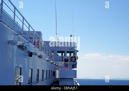 Griechische Inseln zwischen Zypern und Athen. Das Bild zeigt die Seite eines großen weißen Schiffes, möglicherweise eines Fähre- oder Frachtschiffes, mit einem blauen Geländer an der Seite. Das Schiff hat mehrere Fenster und einen kleinen, möglicherweise roten Rettungsschirm, der am Geländer hängt. Auf der Schiffsseite sind zwei hohe Antennen zu sehen, und ein kleiner, möglicherweise roter Rettungsschirm ist ebenfalls in der Nähe des Geländes zu sehen. Das Schiff befindet sich auf dem offenen Wasser, mit einem klaren blauen Himmel und einigen entfernten Formationen im Hintergrund. Das Auge und die Phantasie eines Künstlers Stockfoto