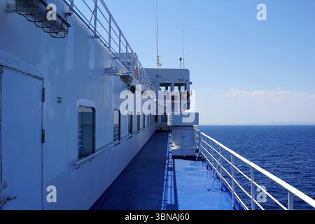 Griechische Inseln zwischen Zypern und Athen. Das Bild zeigt das Deck einer Fähre, in einer makellosen weißen Farbe. Das Deck ist von einem stabilen Geländer umgeben und hat einen blauen Boden. Auf der linken Seite des Bildes ist eine weiße Tür zu sehen. Die Perspektive ist von der Seite der Fähre aus, mit Blick auf den Horizont. Der Himmel ist klar blau, und der Ozean erstreckt sich bis zum Horizont. Die Fähre hat mehrere Fenster entlang der Seite, und eine kleine, weiße, möglicherweise aus Metall bestehende Struktur ist in der Nähe des Geländes zu sehen. Das Auge und die Phantasie eines Künstlers Stockfoto