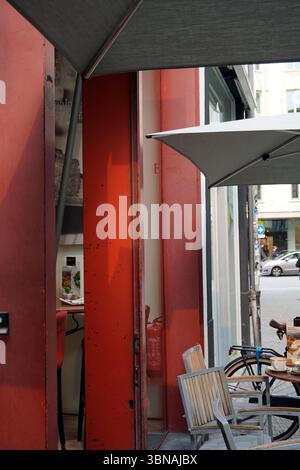 Antwerpen, zweitgrößte belgische Stadt, liegt an der Schelde in Belgien. Eine lebhafte Straßenszene. Der Schwerpunkt ist eine rote Schaufensterfront mit einer leicht offenen Tür, die einen Blick auf das Innere freigibt. Auf der rechten Seite des Rahmens befindet sich ein grauer Regenschirm, der Schatten bietet. Ein Fahrrad steht vor der Ladenfront, und ein kleiner Tisch mit rotem Stuhl befindet sich in der Nähe. Die Storefront hat ein leicht abgenutztes oder veraltetes Aussehen mit sichtbaren Kratzern und Markierungen an der roten Tür. An der Wand neben der Tür ist ein Feuerlöscher zu sehen. Der Bereich vor der Ladenfront ist Stockfoto